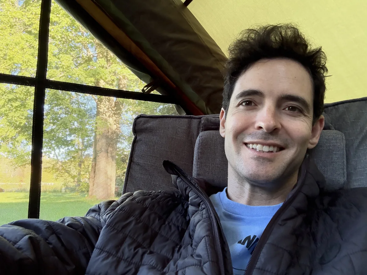 Russell, founder of Camp Komorebi, smiling in a screened tent with trees and afternoon sun behind him